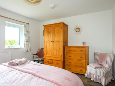 A bedroom with a wooden wardrobe and chest of drawers a bed with pink bedding a floral curtain a striped chair with folded blankets and a small wall mirror at Bwthyn Y Swnt in Moelfre