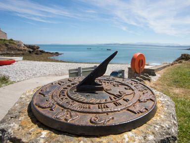 A sundial on a stone base near a pebble beach with boats and a lifebuoy at Bwthyn Y Swnt in Moelfre