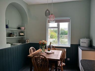 A dining area with wooden table and chairs flowers and wine on table near window with shelves on wall at Secret Garden 34C Clarence Street Dartmouth