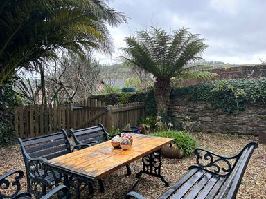 An outdoor garden area with a wooden table and black metal chairs surrounded by plants and a stone wall at Secret Garden 34C Clarence Street Dartmouth