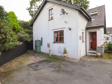 An exterior view of a white cottage with a brown door and windows surrounded by greenery at Cherry Tree Cottage in Windermere