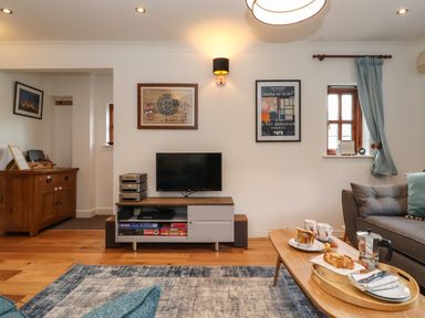 A living room with a wall-mounted television a wooden sideboard a coffee table with breakfast items and a sofa with cushions at Cherry Tree Cottage in Windermere