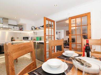A dining area with wooden chairs and table set with plates bowls wine bottle and glasses next to a kitchen with stainless steel appliances at Cherry Tree Cottage in Windermere