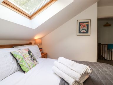 A bedroom with a slanted skylight window a wooden bed with a white pillow and a map-themed cushion a bedside table lamp and folded white towels on a gray blanket at Cherry Tree Cottage in Windermere