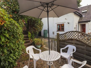An outdoor patio with a white table, four white plastic chairs, and a large umbrella near a white house and wooden fence at Cherry Tree Cottage in Windermere