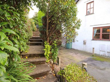 An outdoor area with stone steps surrounded by greenery and a paved section next to a white building with windows at Cherry Tree Cottage in Windermere
