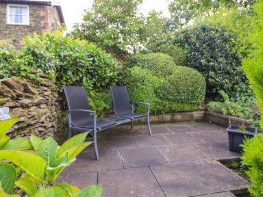 An outdoor patio area with two connected chairs surrounded by bushes and plants at Cherry Tree Cottage in Windermere