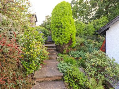 A garden path with stone steps surrounded by various green plants and bushes near a white building at Cherry Tree Cottage in Windermere