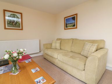 A living room with a beige sofa with two cushions two framed pictures on the wall and a wooden coffee table with a vase of flowers at Hunstone Barn in South Molton