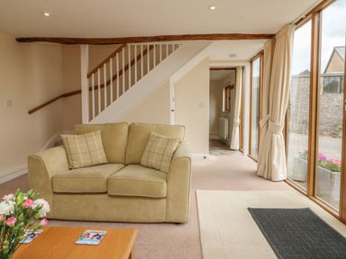A living room with a beige sofa, two plaid cushions, a wooden coffee table with flowers, stairs with a white railing, large windows with curtains, and a doormat at Hunstone Barn South Molton