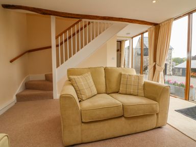 A beige sofa with two plaid cushions in a room with carpeted stairs and large windows with curtains at Hunstone Barn in South Molton