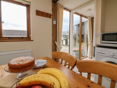 A kitchen with wooden table and chairs a cake and fruit bowl on the table a microwave and washing machine near a sliding glass door at Hunstone Barn in South Molton
