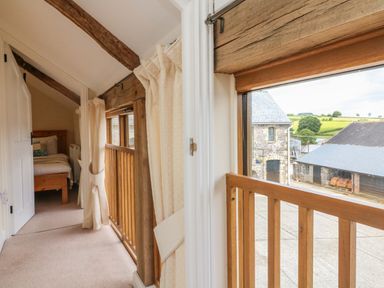 A hallway with windows and curtains overlooking an outdoor farm area with stone buildings at Hunstone Barn in South Molton