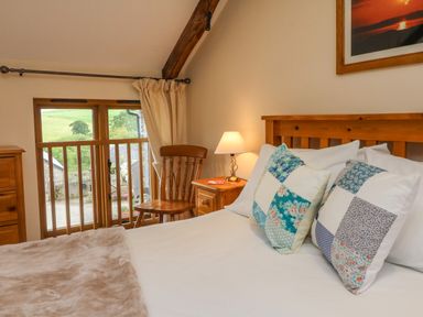 A bedroom with a wooden bed frame two pillows a wooden chair a nightstand with a lamp and a window with curtains at Hunstone Barn in South Molton