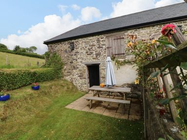 A stone barn with a wooden picnic table and umbrella on a patio with grass and flowers at Hunstone Barn in South Molton