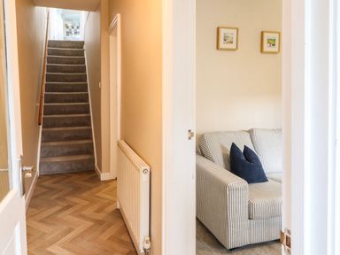 A hallway with carpeted stairs and a radiator next to a room with a striped sofa and two framed pictures on the wall at Bay Tree Cottage Keswick
