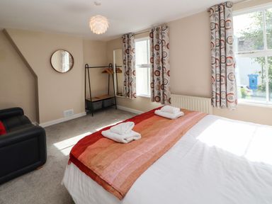 A bedroom with a bed, folded towels, a sofa, a clothes rack with hangers, a round wall mirror, and patterned curtains at Bay Tree Cottage in Keswick