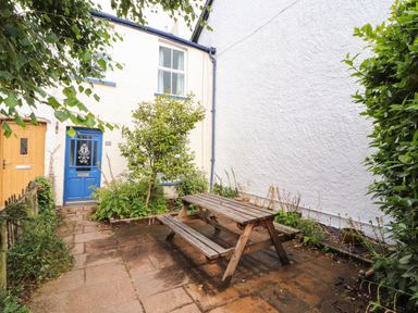 A small outdoor patio area with a wooden picnic table and benches outside a house with a blue door and window frames at Bay Tree Cottage Keswick