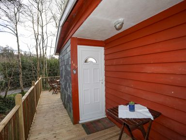 A wooden balcony with a white door red siding a small table with a plant and outdoor chairs at Treetops in Caernarfon