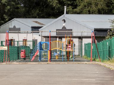 A fenced playground with swings slides and climbing structures near industrial buildings at Treetops in Caernarfon