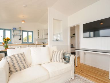 A living room with a white sofa and striped pillows a TV mounted on the wall and a kitchen with blue window frames and wooden countertop at Solana Whitsand Bay