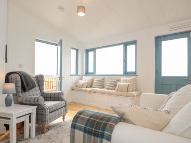 A living room with a plaid armchair a window seat with cushions and a white side table with a lamp at Solana Whitsand Bay