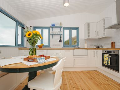 A kitchen with a wooden round table white chairs flowers croissants and fruit bowl near blue windows at Solana in Whitsand Bay
