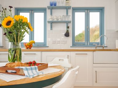 A kitchen with blue window frames a wooden countertop white cabinets a vase with sunflowers and a tray with croissants and strawberries at Solana Whitsand Bay