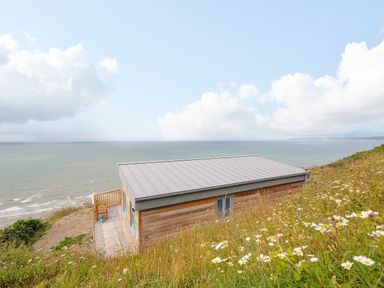 A wooden cabin with a metal roof on a grassy hill overlooking the ocean at Solana in Whitsand Bay