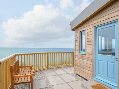 A wooden bench and railing on a patio overlooking the ocean next to a wooden structure with a blue door and window at Solana in Whitsand Bay
