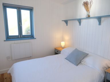 A bedroom with a bed a side table with a lamp a window and a wall shelf with a vase at Solana in Whitsand Bay