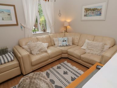 A living room with a cream leather corner sofa and cushions beside a window with white curtains and framed pictures at Albert's Den in Falmouth