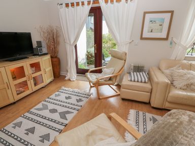 A living room with beige chairs a sofa a wooden cabinet a television white curtains and patterned rugs at Alberts Den in Falmouth