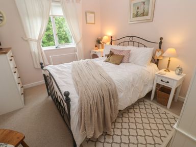 A bedroom with a bed with a white duvet and pillows a beige throw blanket bedside tables with lamps and an alarm clock a dresser a window with curtains and a patterned rug at Alberts Den in Falmouth