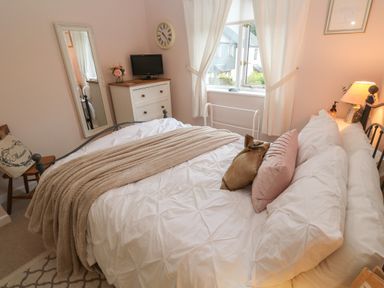 A bedroom with a bed with white bedding and pillows a chest of drawers with a small tv a wall clock a full-length mirror and a window with curtains at Albert's Den in Falmouth