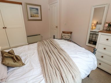A bedroom with a bed covered by a beige throw and white bedding a wooden dresser with a mirror and flowers a wardrobe and a wall picture at Alberts Den in Falmouth