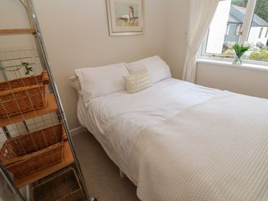 A bedroom with a bed with white bedding and a small pillow near a window with white curtains and a vase of flowers on the windowsill at Albert's Den in Falmouth