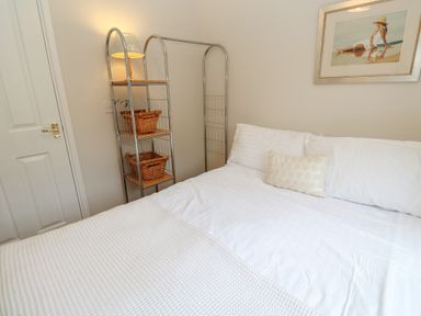 A bedroom with a bed white bedding a shelving unit with baskets and a lamp a closed door and a framed artwork on the wall at Alberts Den in Falmouth