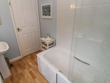 A bathroom with a bathtub a glass shower screen a towel hanging on the tub a sink a small cabinet with baskets and toiletries a framed picture on the wall at Albert's Den in Falmouth