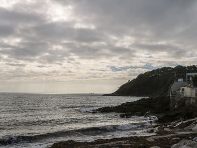 A rocky coastline with waves and a hill covered in trees beside buildings at Alberts Den in Falmouth