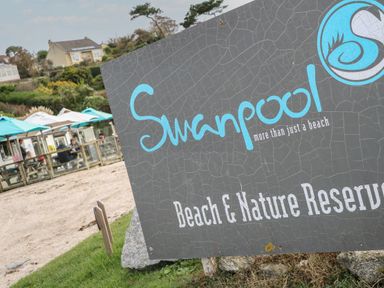A sign for Swanpool Beach and Nature Reserve with people sitting under blue umbrellas near the beach at Albert's Den in Falmouth