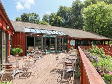 An outdoor wooden deck with tables and chairs in front of a building labeled Pendra Loweth Bar surrounded by trees and plants at Albert's Den in Falmouth