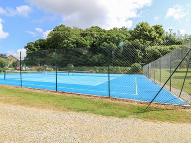 An outdoor tennis court with blue surface surrounded by a chain link fence and trees in the background at Alberts Den in Falmouth