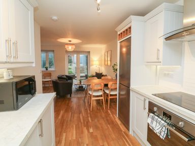 A kitchen with white cabinets and appliances opening into a dining area with a wooden table and chairs a sitting area with black chairs and glass doors at Reubens Cottage in Fremington Devon