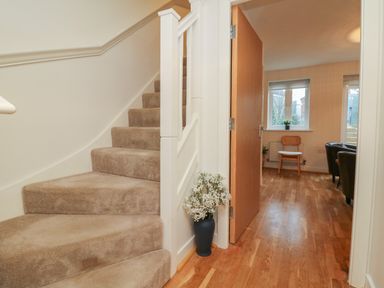 A hallway with carpeted stairs and a vase with flowers next to an open door leading to a room with wooden floor and chair at Reubens Cottage in Fremington Devon