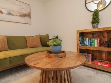 A seating area with a green sofa and checked pillows a wooden round table with a plant and a wooden bookshelf with books and games at Reubens Cottage in Fremington Devon