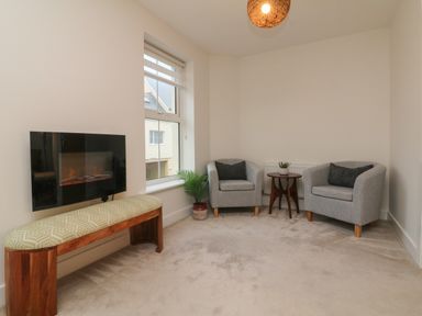 A sitting room with two gray armchairs a small wooden table bench and wall-mounted electric fireplace at Reubens Cottage in Fremington Devon