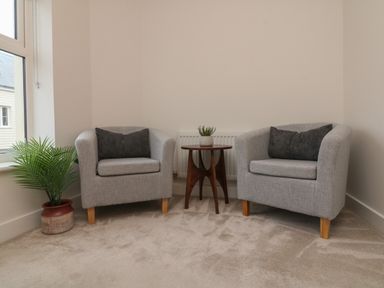 Two grey armchairs with dark cushions separated by a wooden side table with a small plant and a potted plant near a window at Reubens Cottage in Fremington Devon