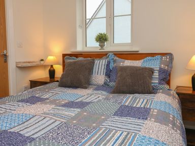 A bedroom with a bed with patterned bedding two brown pillows two bedside tables with lamps and a window with a small plant on the sill at Reubens Cottage in Fremington Devon