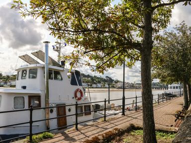 A riverside pathway with trees benches and boats docked along the water at Reubens Cottage in Fremington Devon